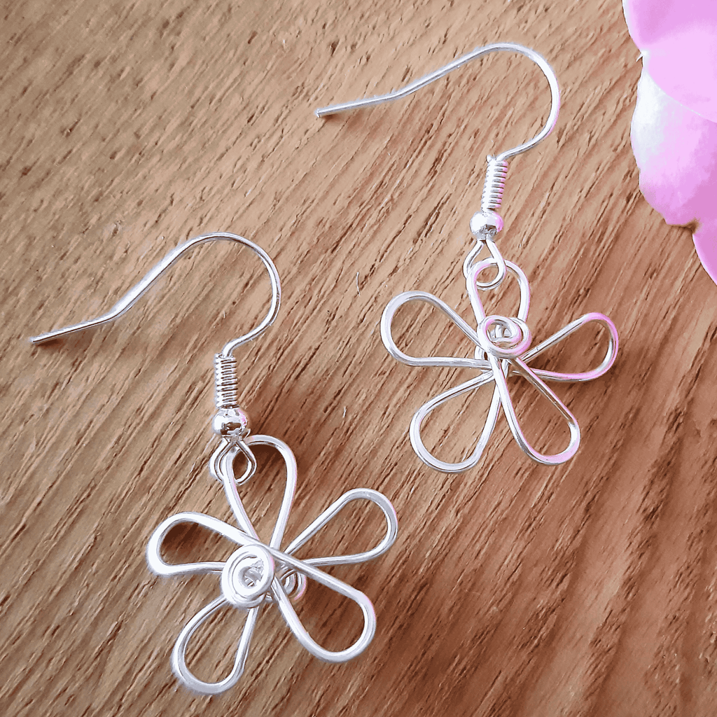Silver flower-shaped earrings on a wooden surface with pink flowers in the background