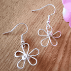 Silver flower-shaped earrings on a wooden surface with pink flowers in the background