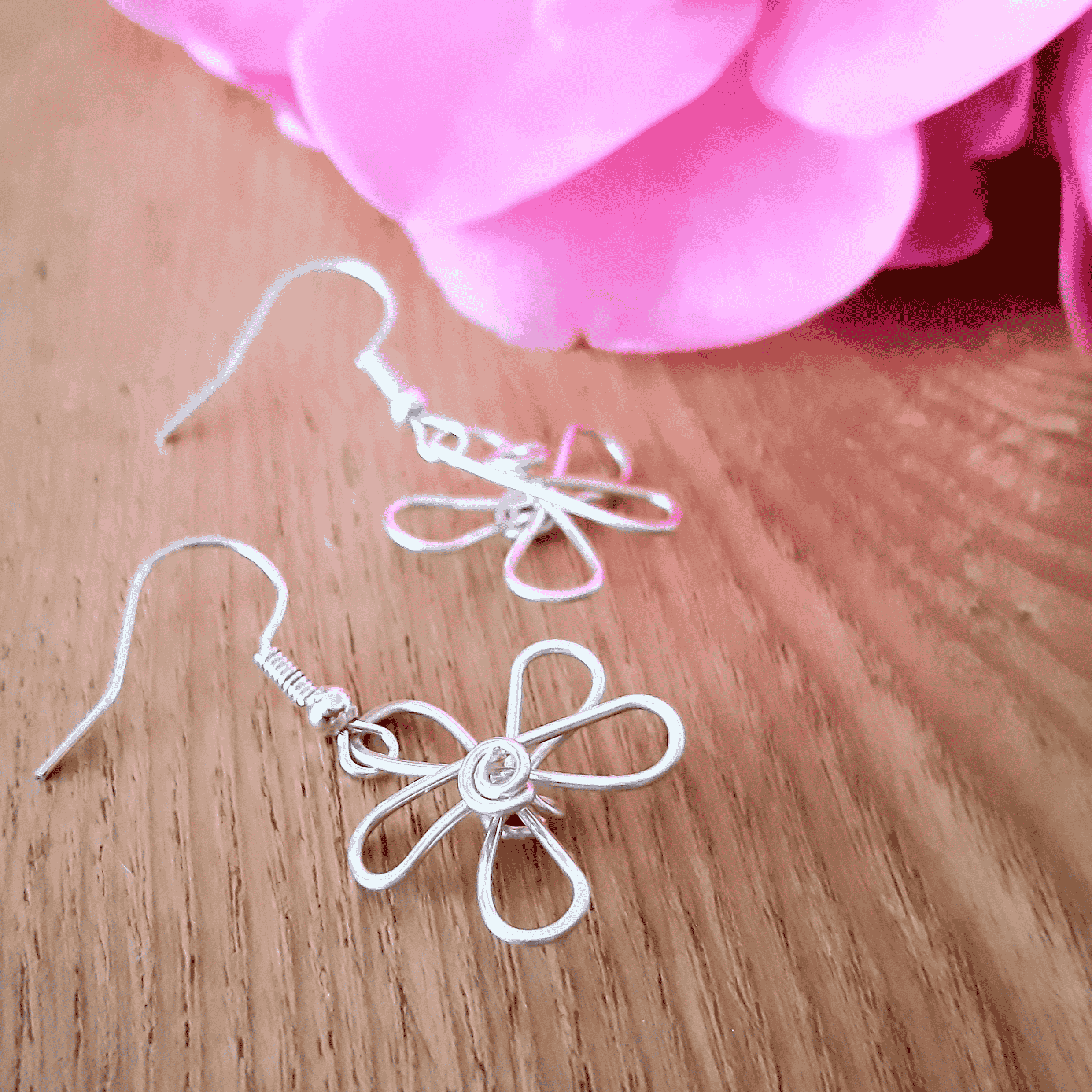 Silver flower-shaped earrings on a wooden surface with pink fabric in the background