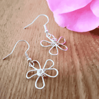 Wire-wrapped flower-shaped earrings on a wooden surface with a pink flower in the background.