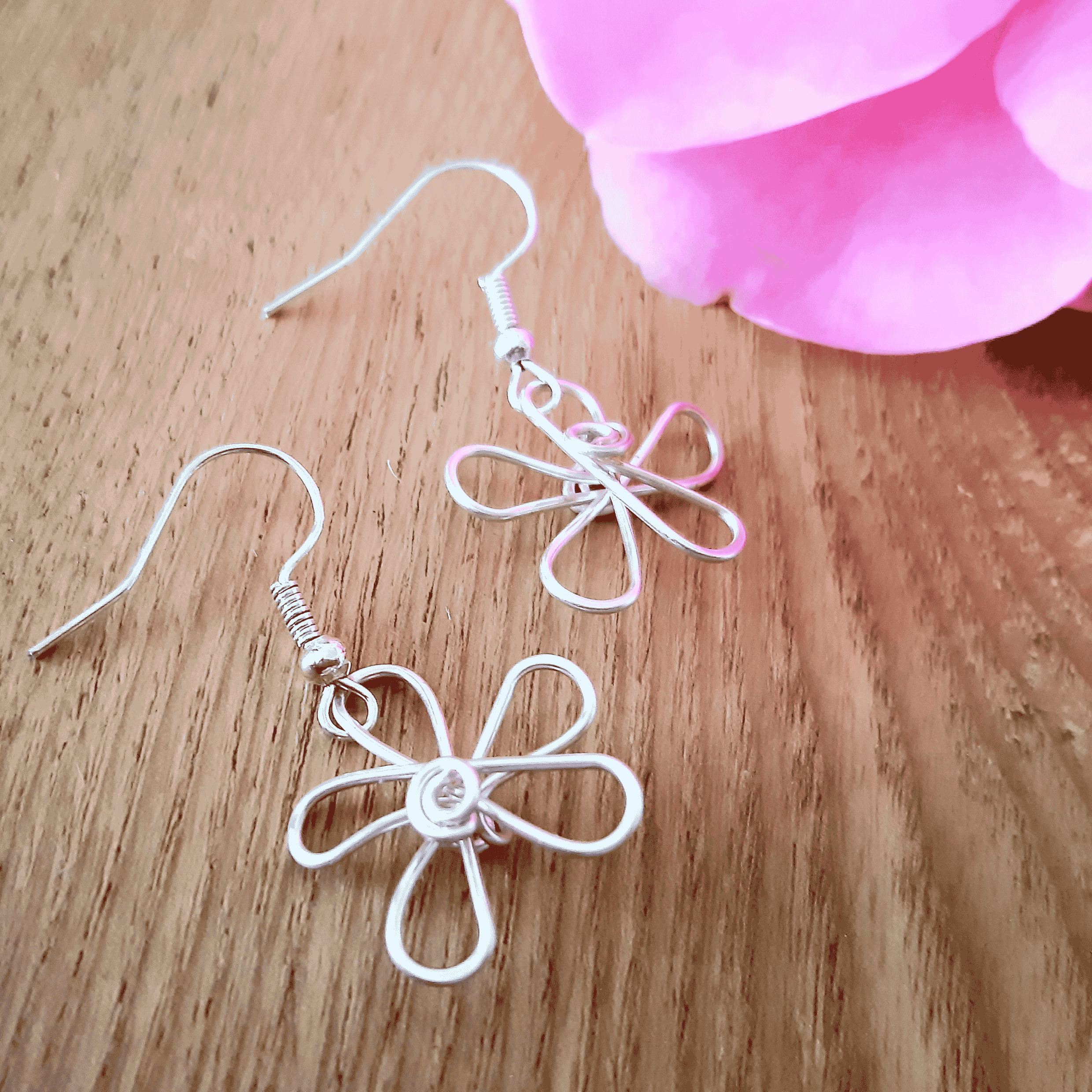 Wire-wrapped flower-shaped earrings on a wooden surface with a pink flower in the background.