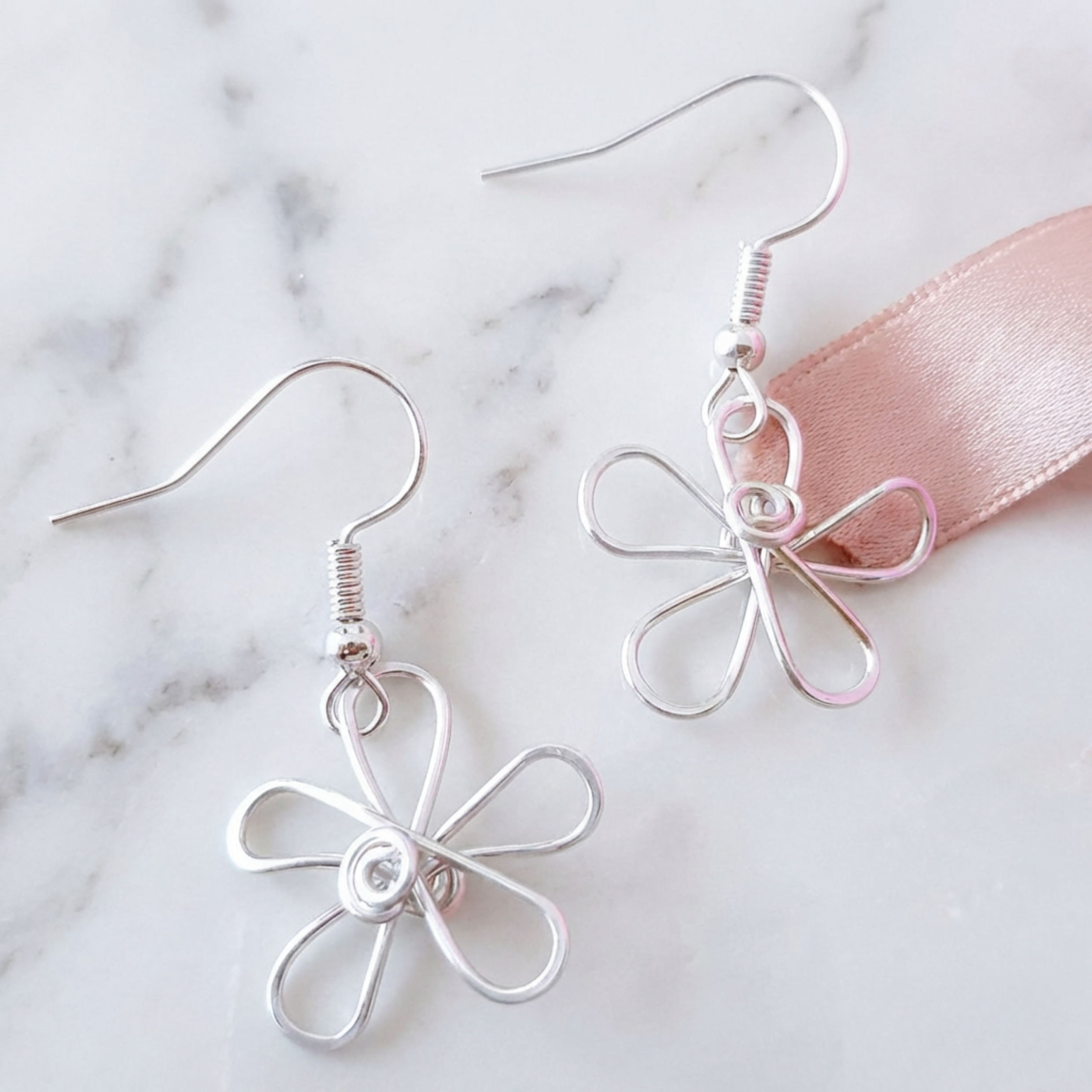 Silver flower-shaped earrings on a marble surface with a pink ribbon.