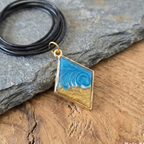 Blue wave pendant on gold chain with black cord on stone and wood background