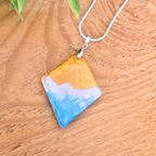 Necklace with a square diamond pendant featuring blue, white, and gold colours in a beach wave effect on a wooden surface.