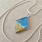 Necklace with a square pendant featuring blue and gold colors on a beige fabric background
