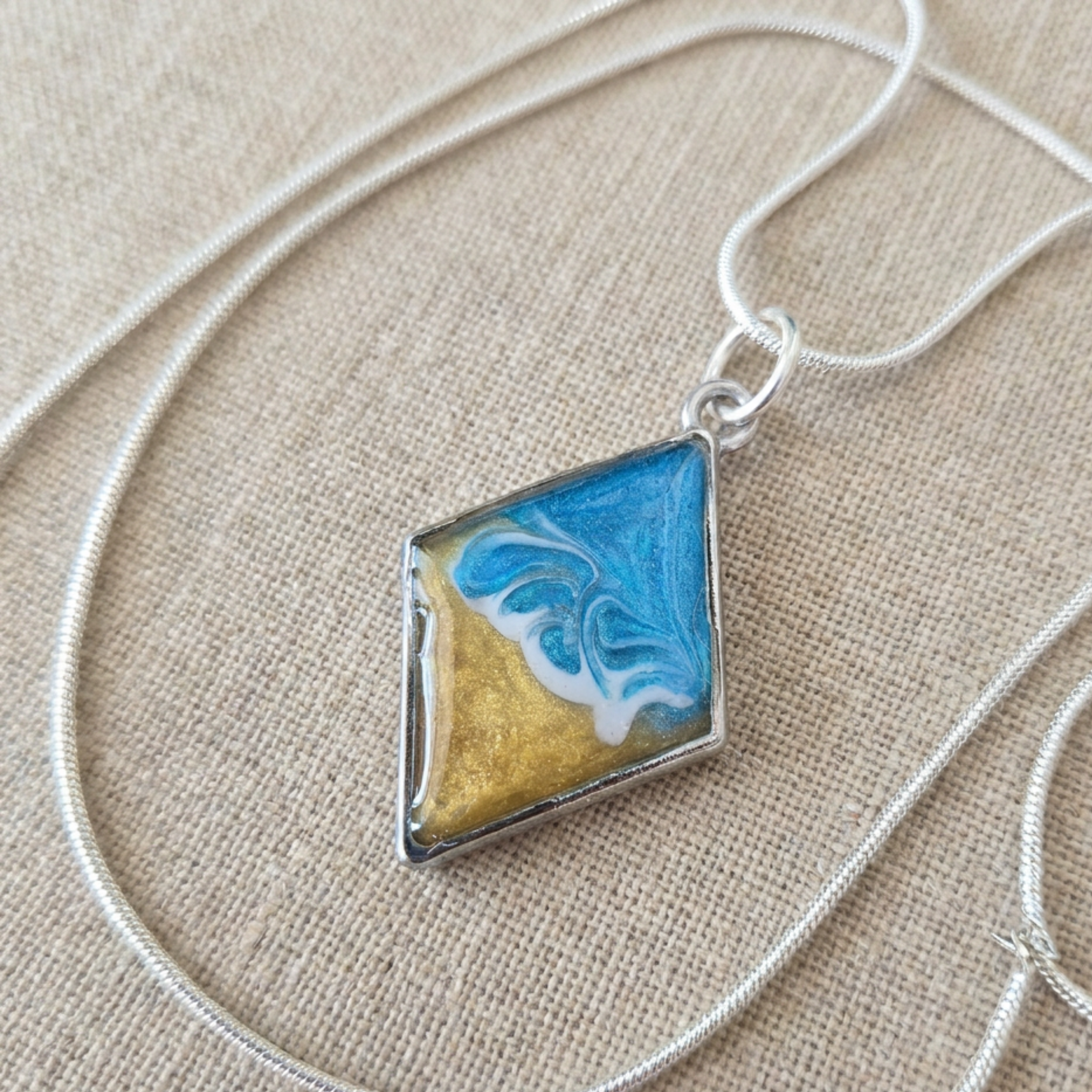 Necklace with a square pendant featuring blue and gold colors on a beige fabric background