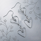 Silver heart-shaped earrings on a textured silver background with leaf patterns.