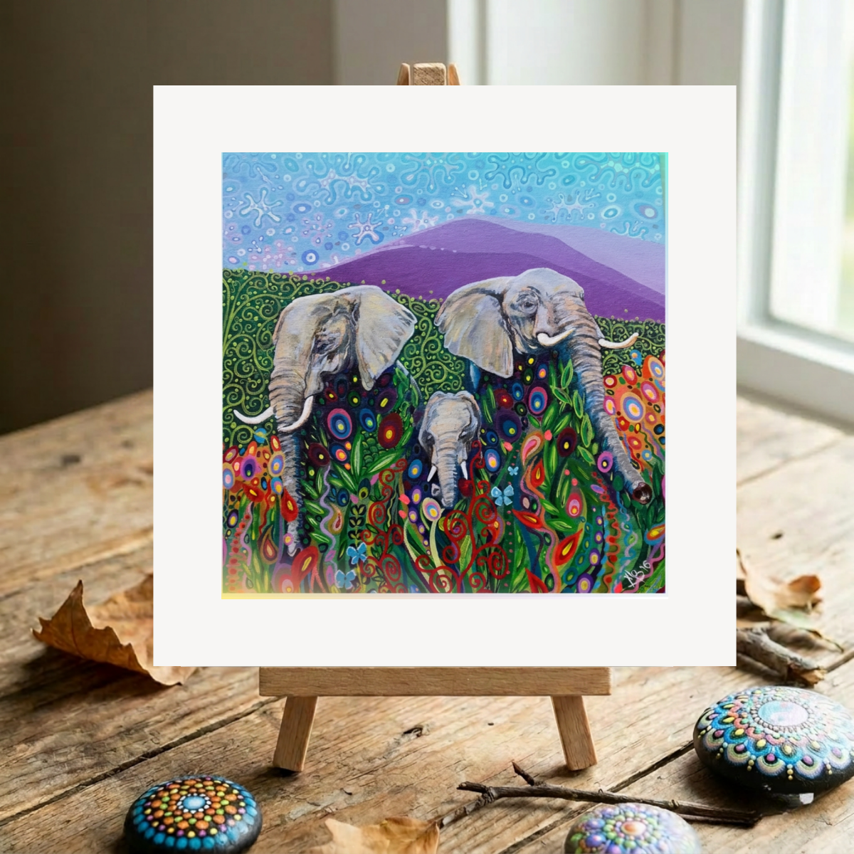 Colourful painting of elephants on a wooden easel with decorative stones and leaves.