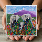 Colourful painting greeting card of three elephants held by a person on a wooden surface