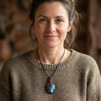 Woman wearing a brown sweater with a necklace featuring a blue pendant, against a blurred background.