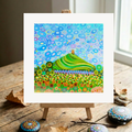 Colourful abstract artwork of Glastonbury Tor on a wooden easel with decorative stones on a wooden surface.