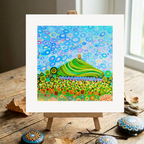 Colourful abstract artwork of Glastonbury Tor on a wooden easel with decorative stones on a wooden surface.
