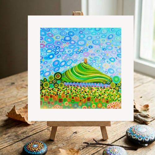 Colourful abstract artwork of Glastonbury Tor on a wooden easel with decorative stones on a wooden surface.