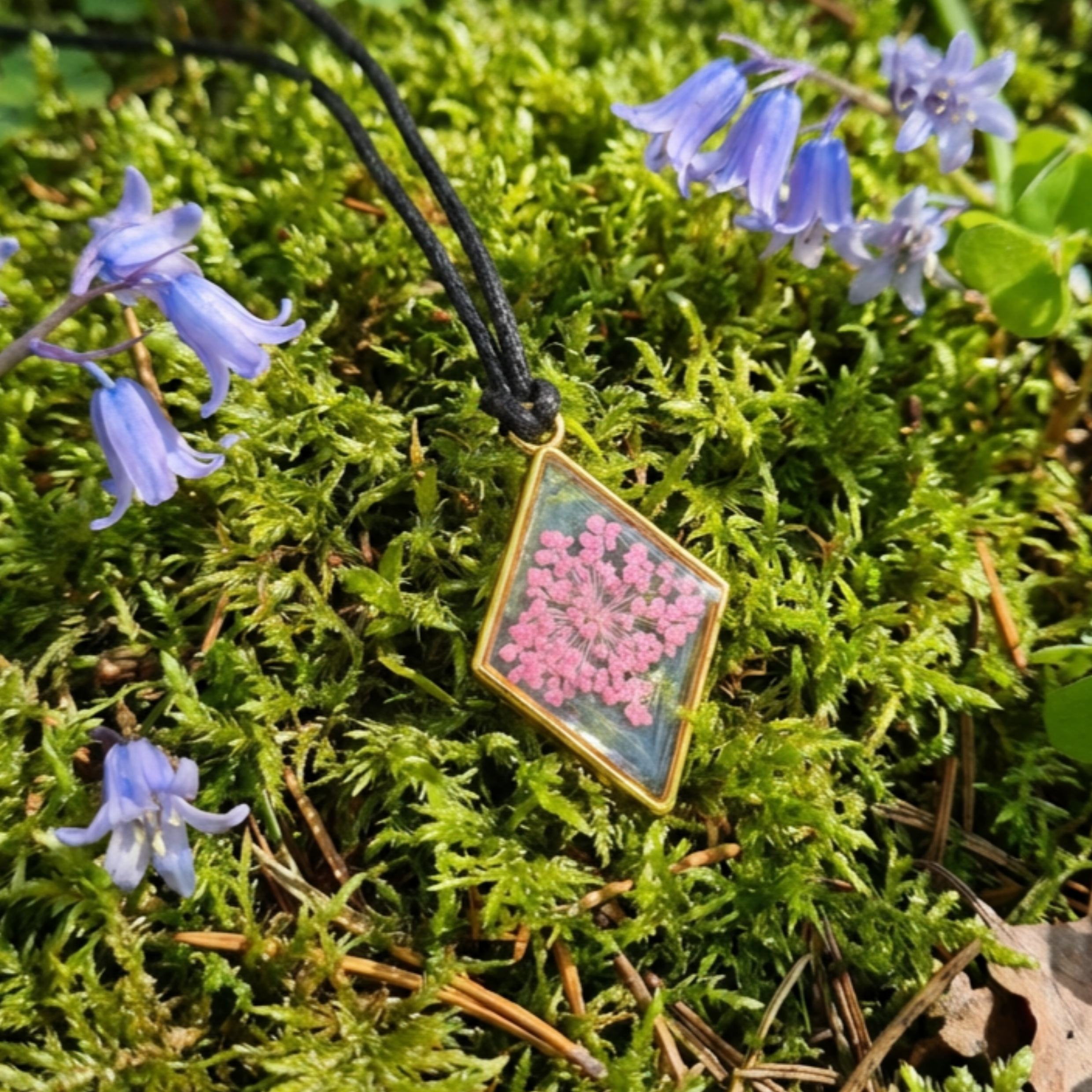 Gold Diamond bezel with clear centre suspending a pink flower pendant on a moss surface