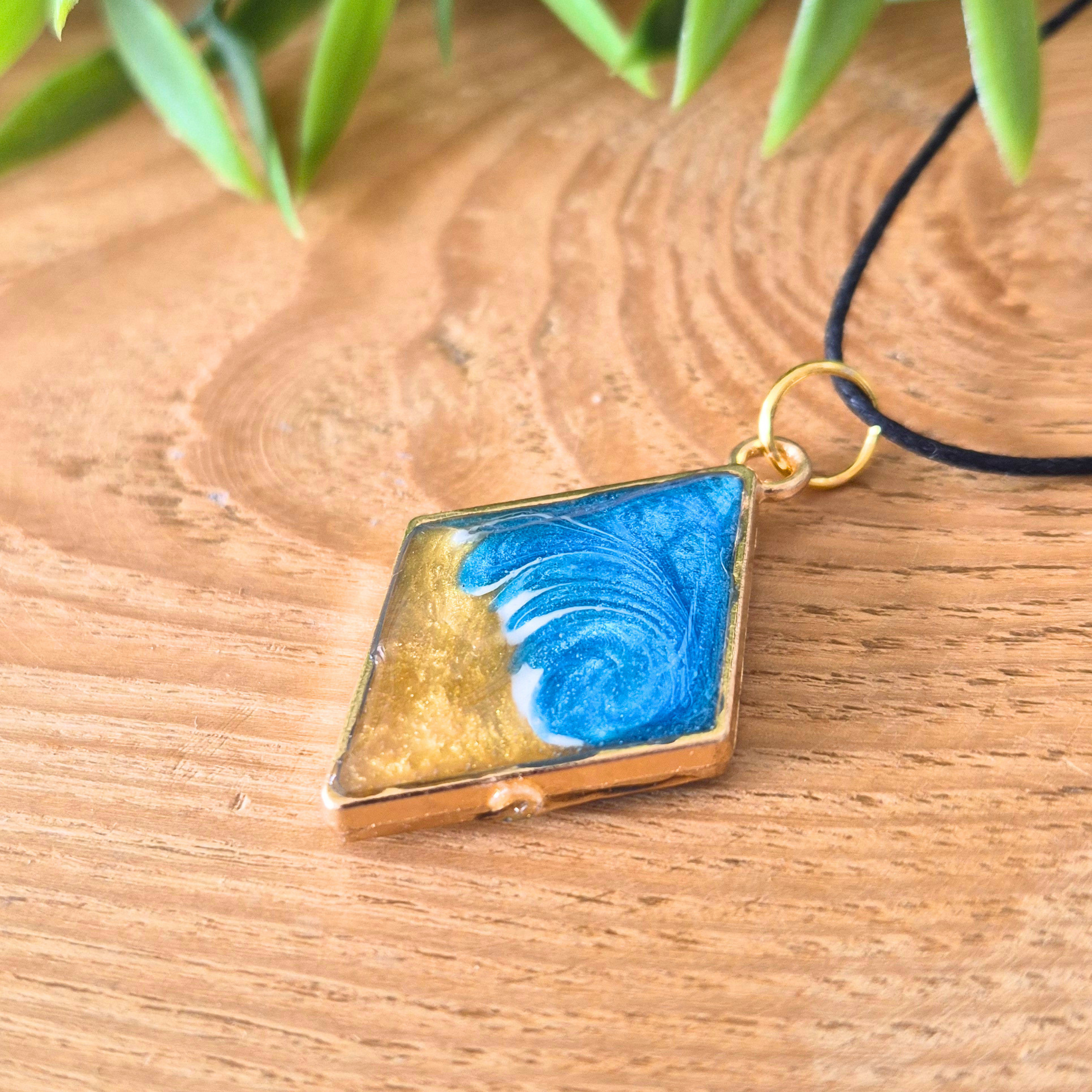 Gold and blue pendant on a wooden surface with green leaves in the background