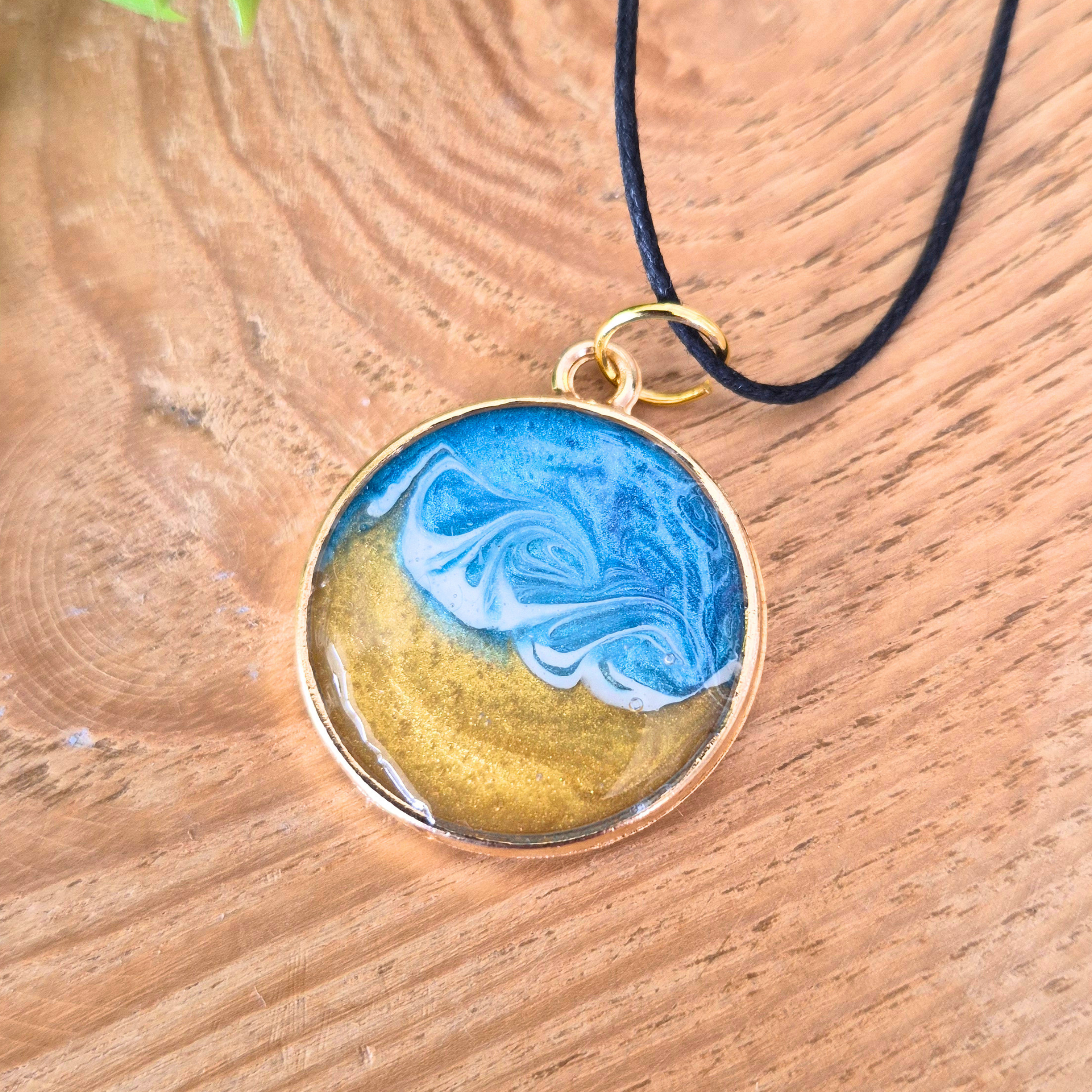 Round pendant with blue and gold design on a black cord, placed on a wooden surface.