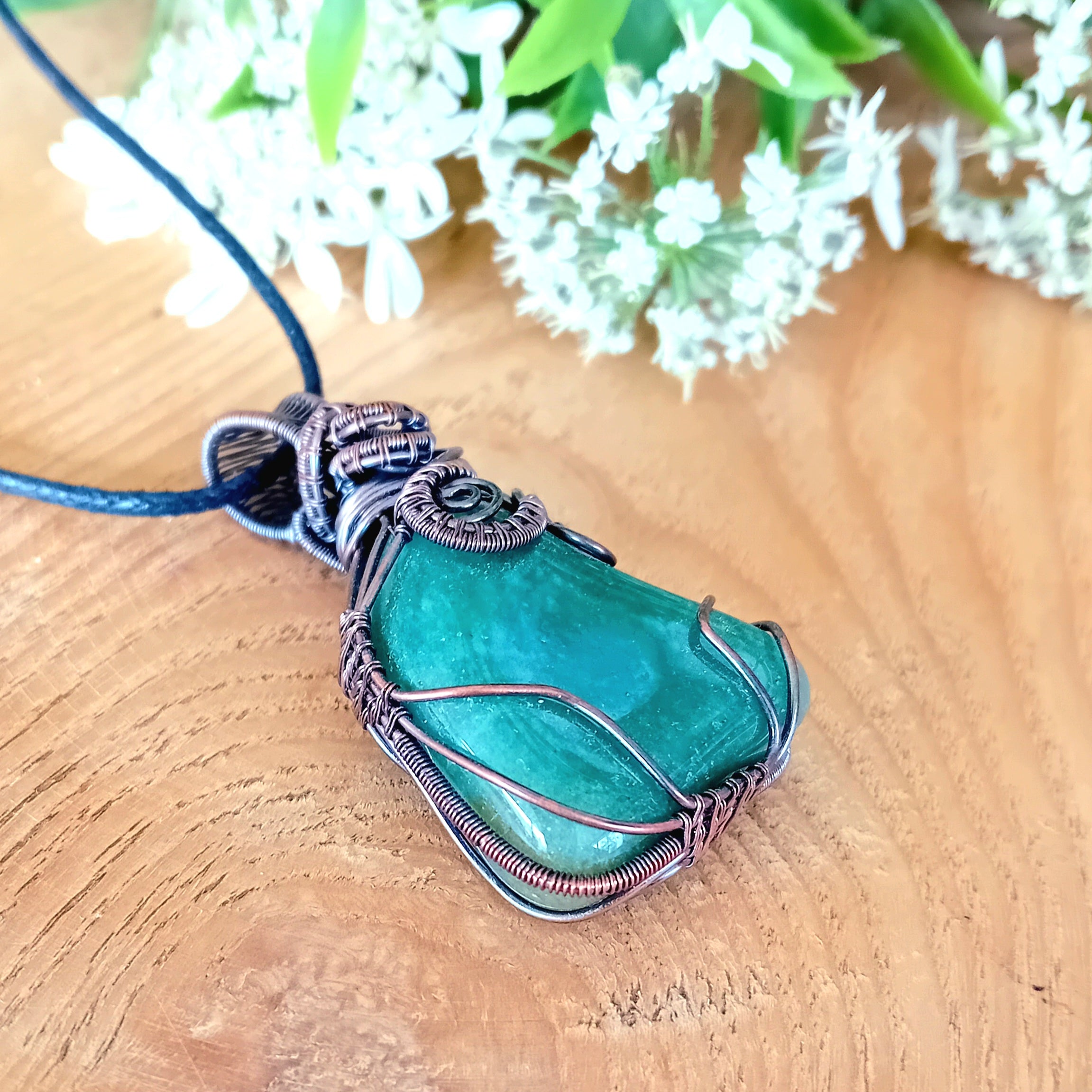 Aquamarine stone pendant with copper wire wrapping on a wooden surface with white flowers in the background