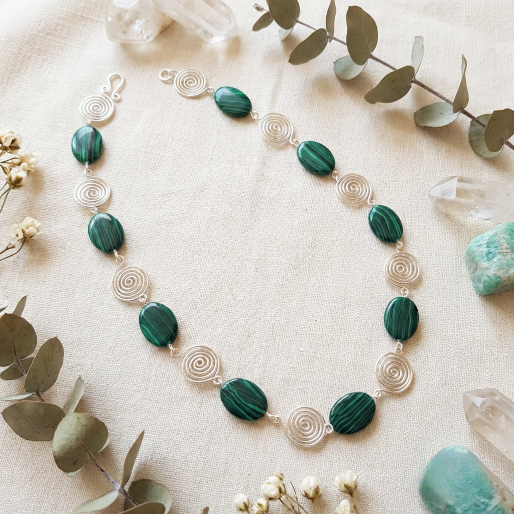 Necklace with green beads and silver spiral accents on a beige surface with eucalyptus leaves and crystals.