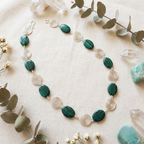 Necklace with green beads and silver spiral accents on a beige surface with eucalyptus leaves and crystals.