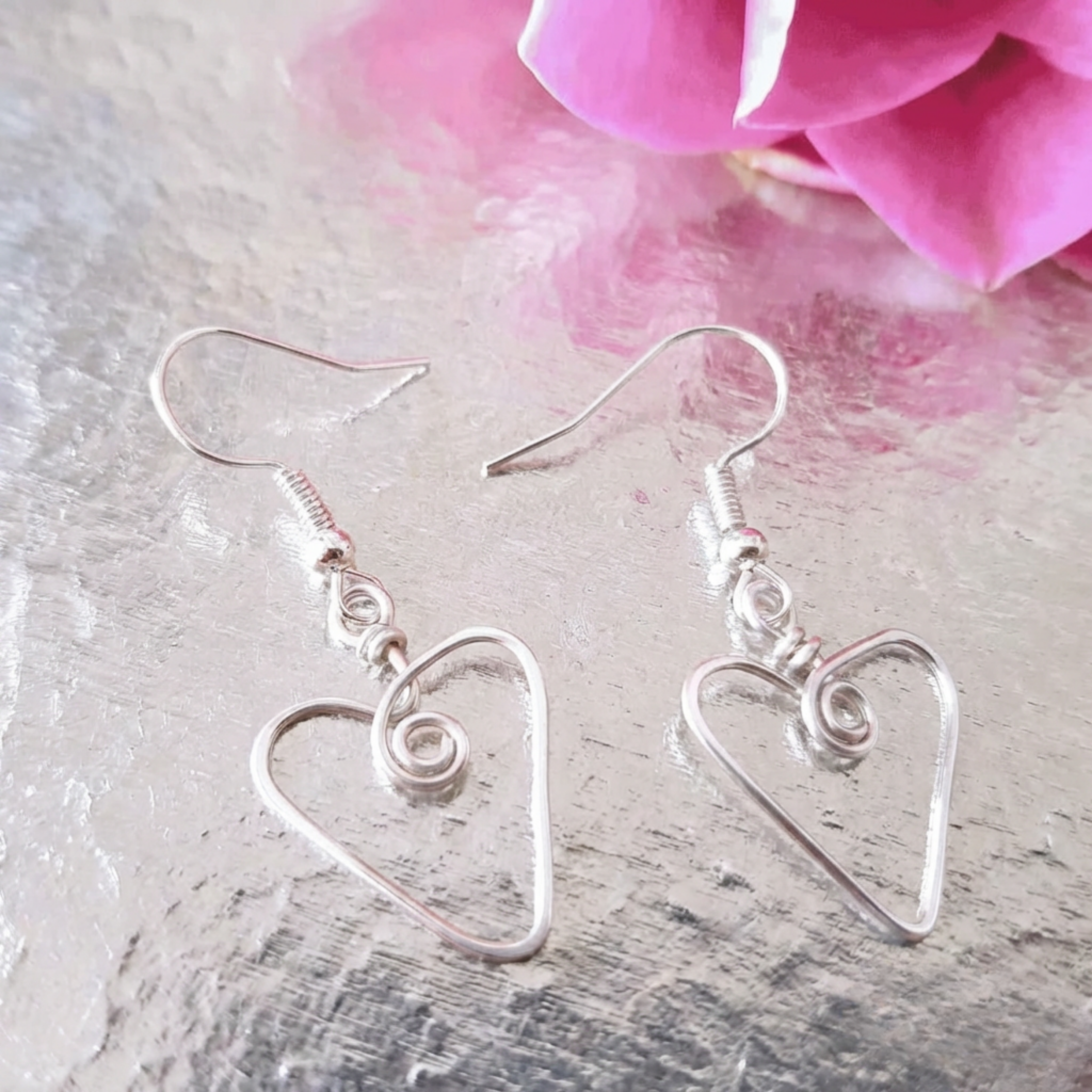 Silver heart-shaped earrings on a textured surface with a pink flower in the background