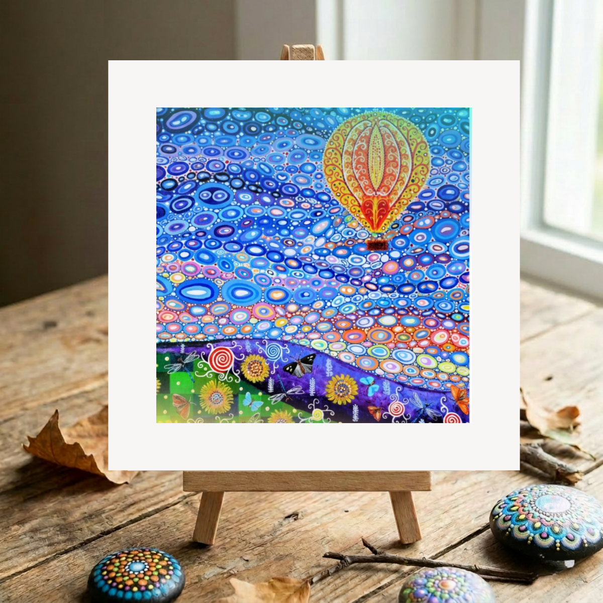 Colourful abstract artwork on a wooden easel with decorative stones on a wooden surface.