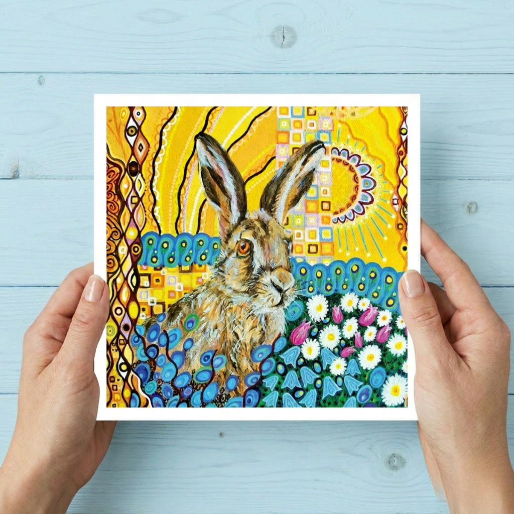 Colourful artwork of a hare held by hands against a light blue wooden background