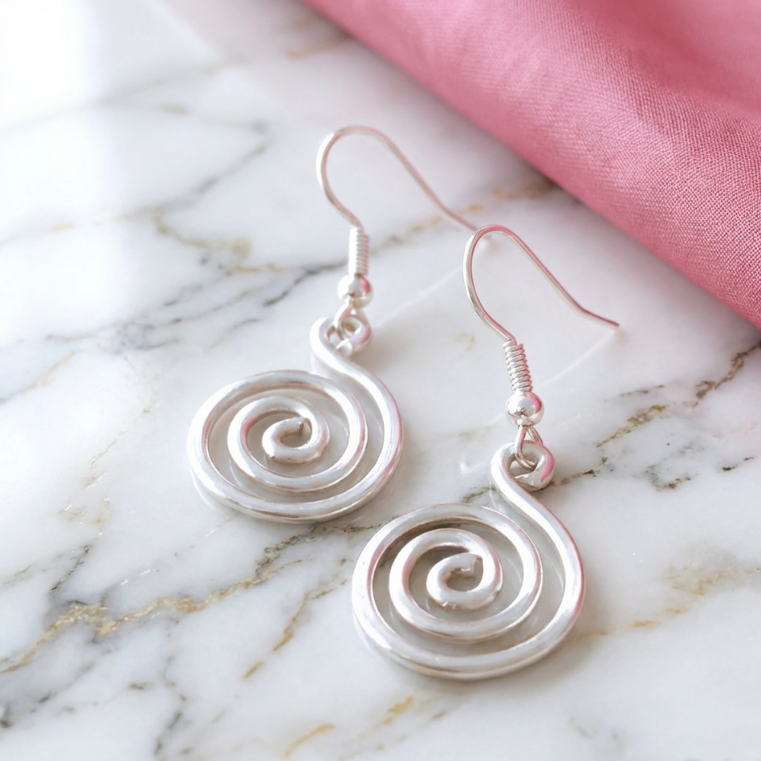 Silver spiral earrings on a marble surface with pink fabric in the background