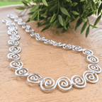 Silver necklace with intricate designs on a wooden surface with green leaves in the background