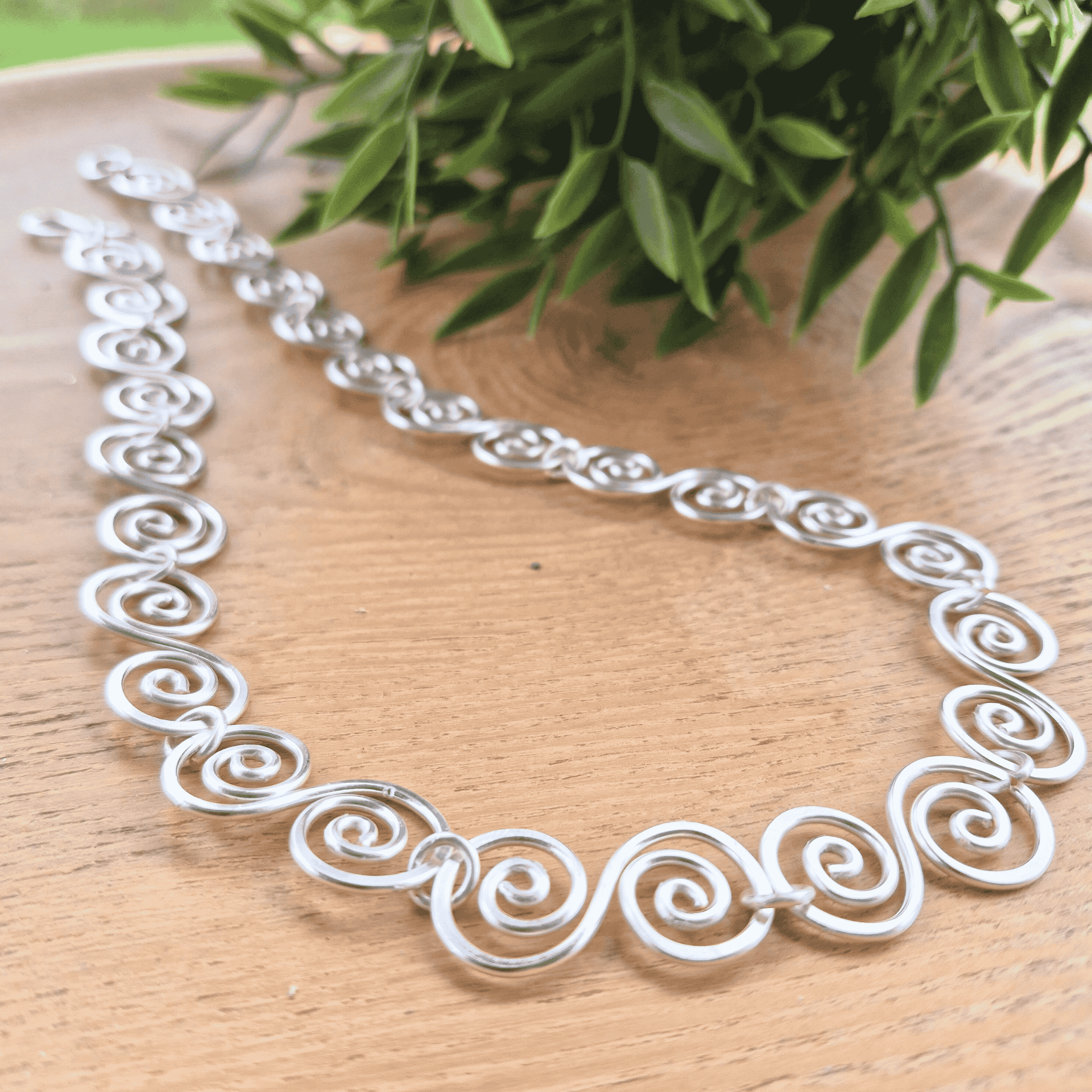 Silver necklace with intricate designs on a wooden surface with green leaves in the background