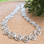 Silver necklace with intricate designs on a wooden surface with greenery in the background