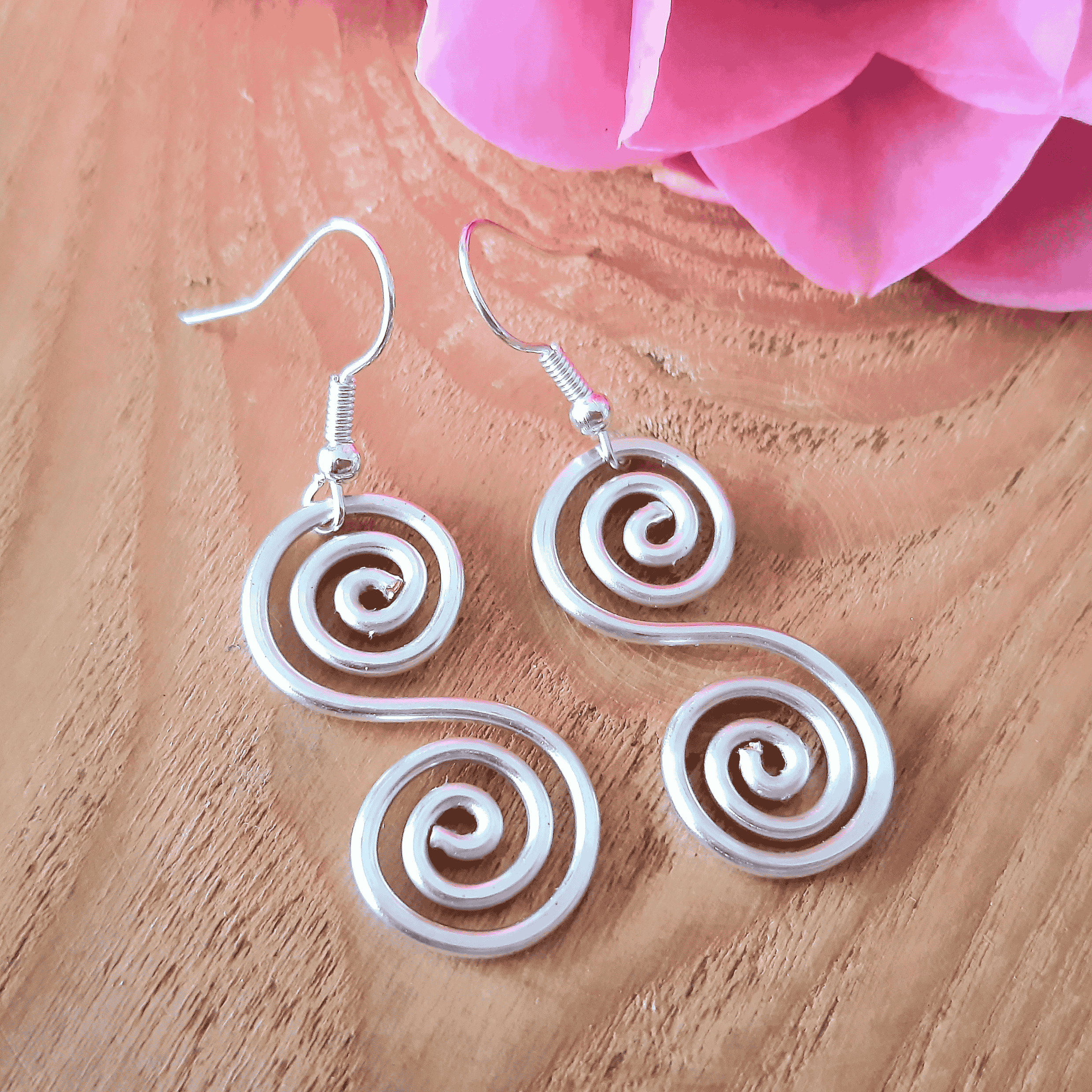 Silver spiral earrings on a wooden surface with pink flowers in the background
