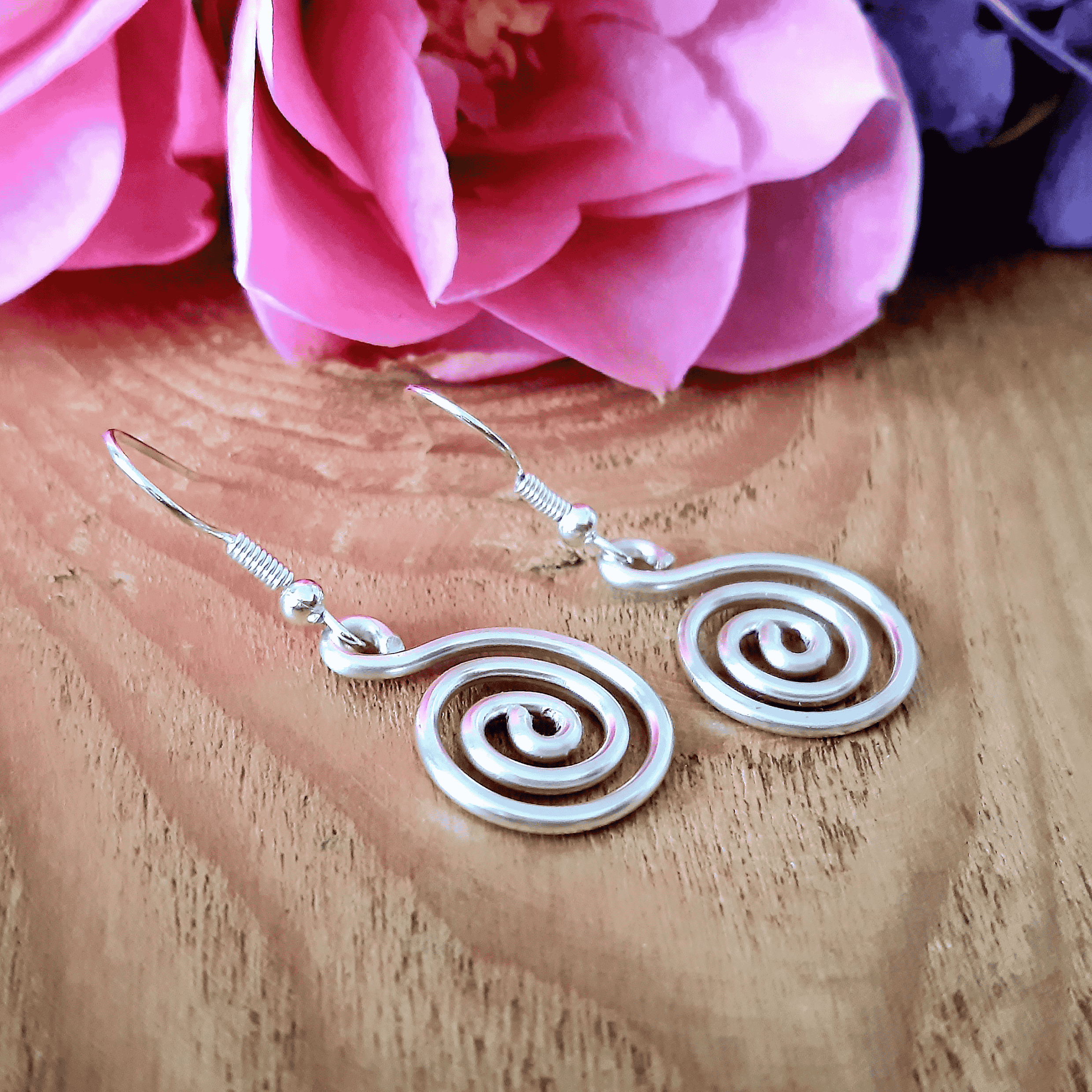 Silver spiral earrings on a wooden surface with pink flowers in the background