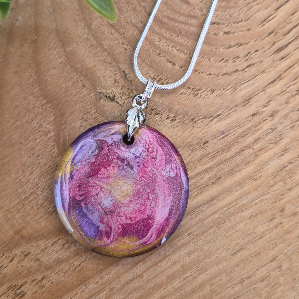 Circular pendant with shimmers of pink purple and gold swirls on a leaf bail and a silver chain on a wooden surface