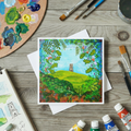 Colourful landscape painting of Glastonbury Tor on a card with art supplies on a wooden surface