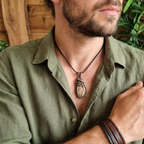 Man wearing a green shirt with a necklace featuring a large pendant, set against a wooden and plant background.