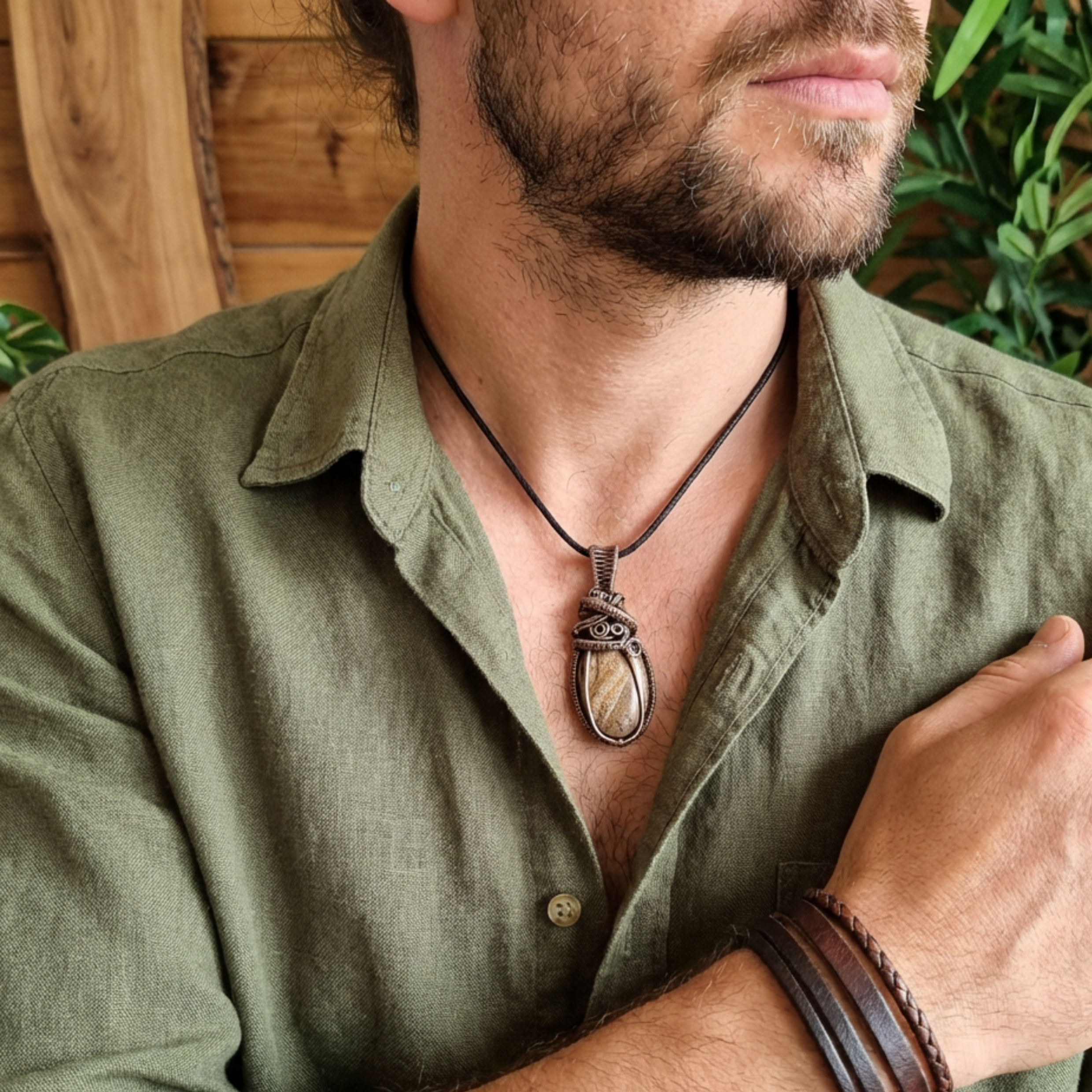 Man wearing a green shirt with a necklace featuring a large pendant, set against a wooden and plant background.