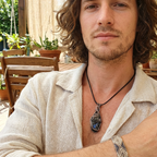 Man with long hair and beard wearing a necklace with a pendant, sitting outdoors.