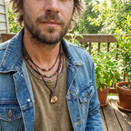 Man wearing a denim jacket and multiple necklaces outdoors on a wooden deck with plants in the background