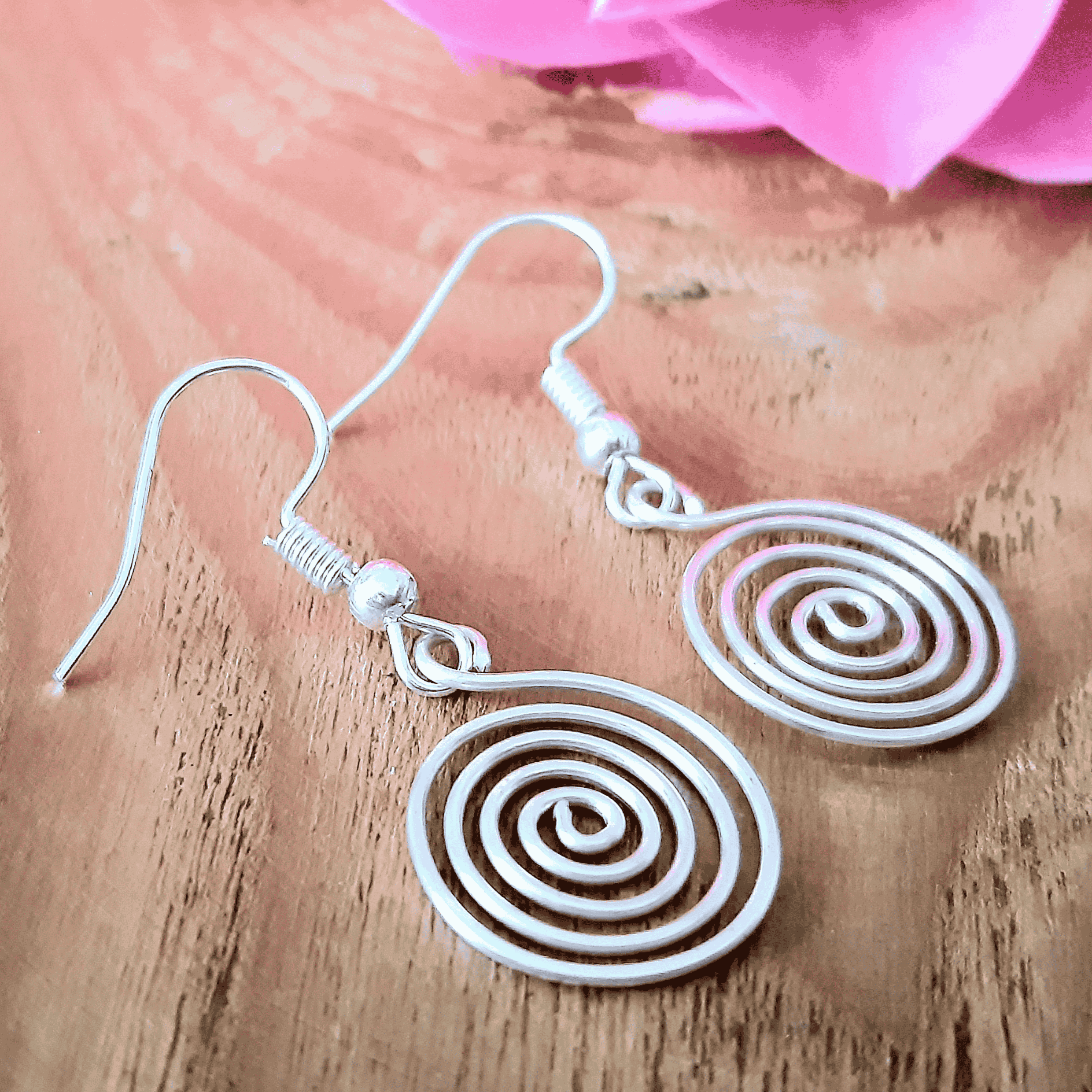 Silver spiral earrings on a wooden surface with a pink flower in the background