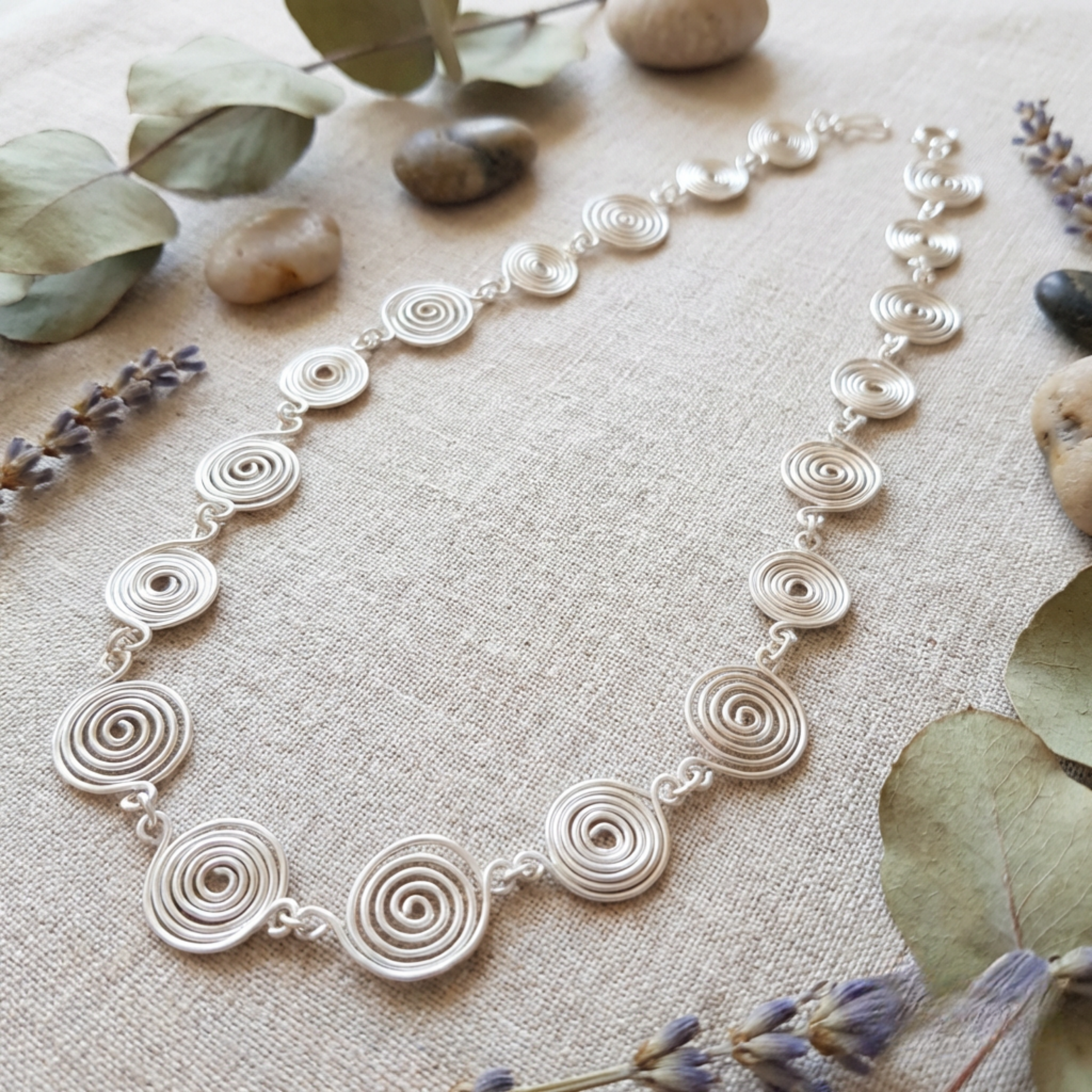 Silver spiral necklace on a textured surface with stones and lavender flowers