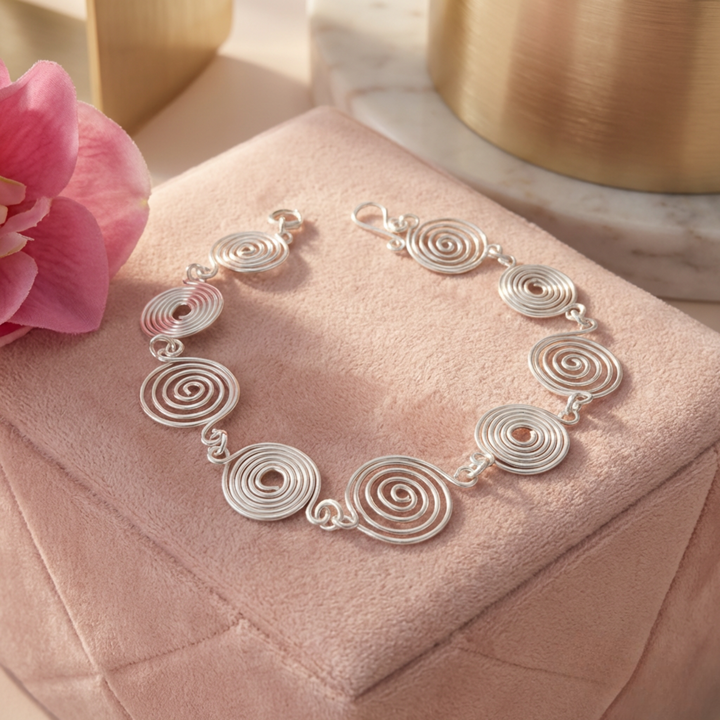 Silver spiral bracelet on a pink cushion with a pink flower and gold object in the background