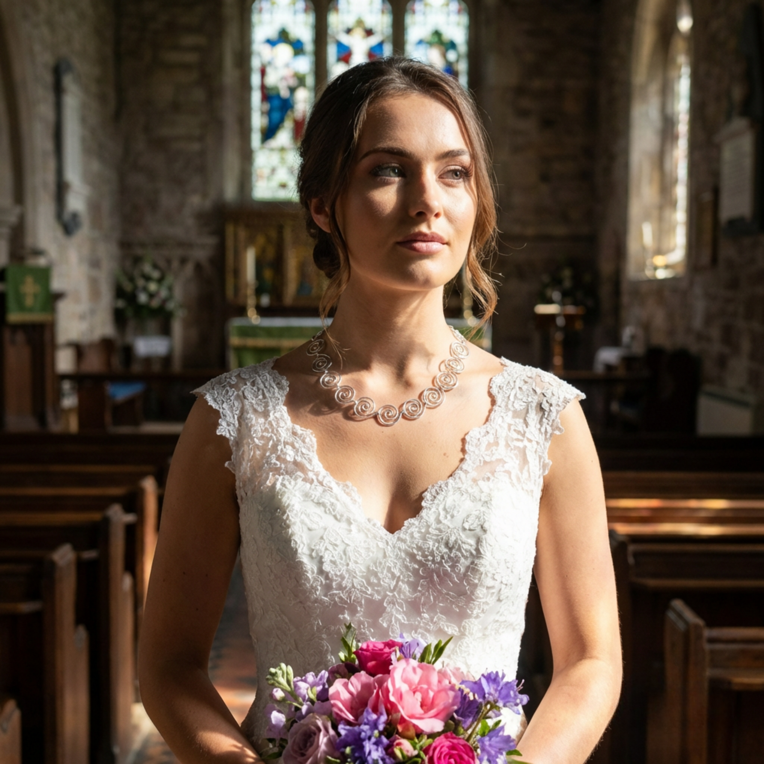 Woman in a white lace dress holding a bouquet of flowers in a church setting wearing a large silver spiral necklace
