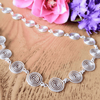 Detailed view of a Silver spiral necklace on a wooden surface with pink and purple flowers.