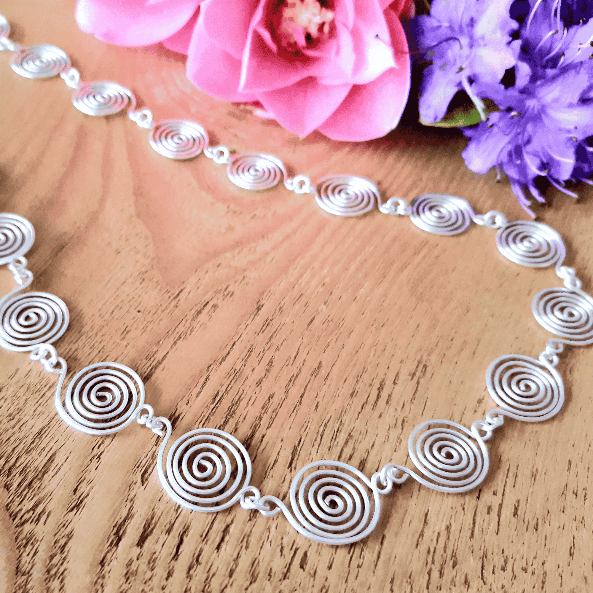 Detailed view of a Silver spiral necklace on a wooden surface with pink and purple flowers.