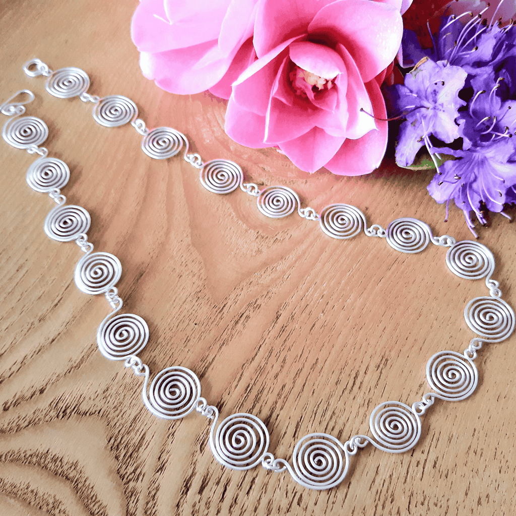Silver necklace with spiral design on a wooden surface with pink and purple flowers.
