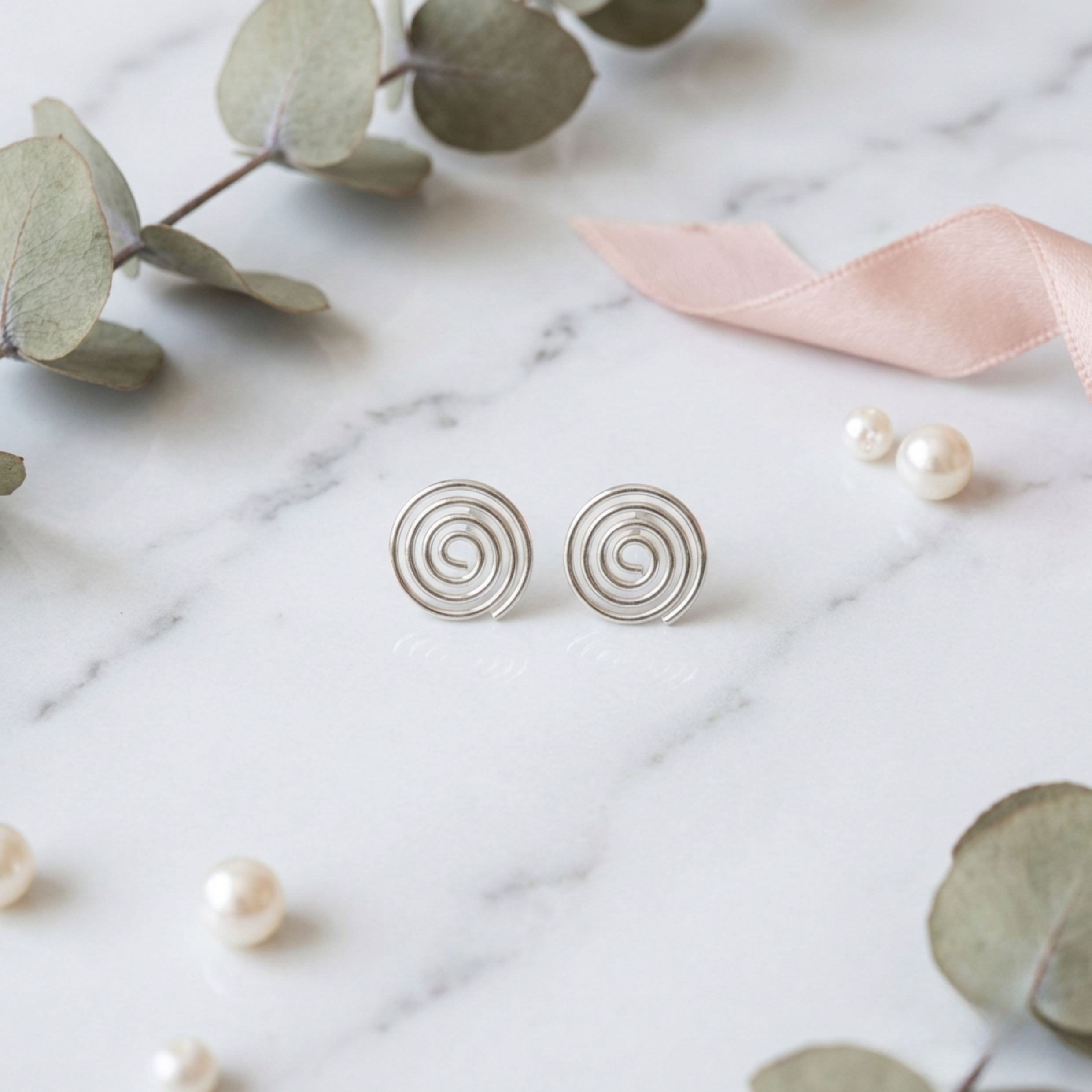 Silver spiral earrings on a marble surface with eucalyptus leaves and a pink ribbon.