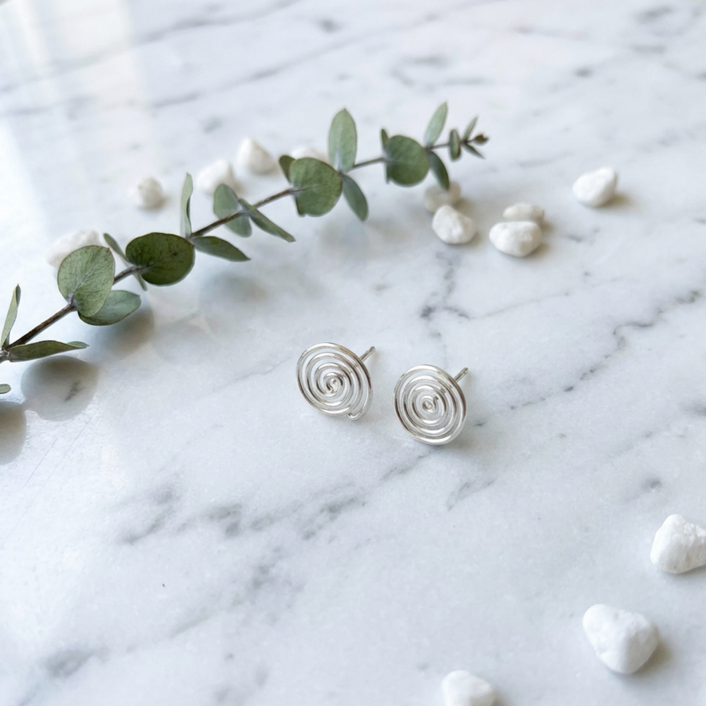 Silver spiral earrings on a marble surface with eucalyptus leaves and small stones.