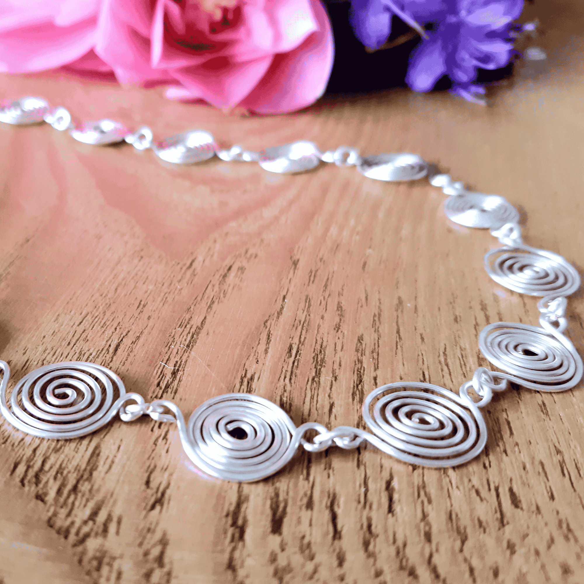 Silver spiral necklace on a wooden surface with flowers in the background