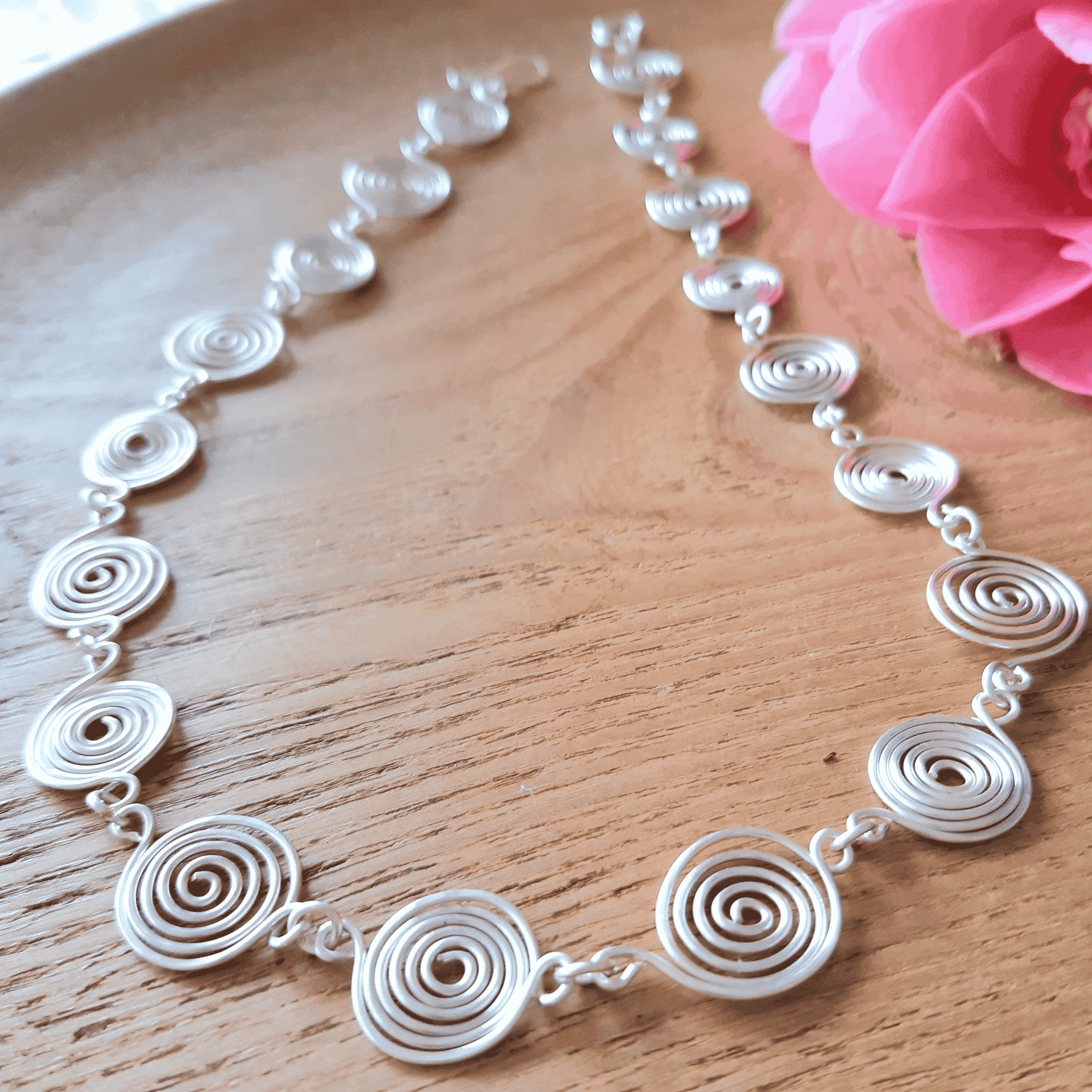 Silver spiral necklace on a wooden surface with a pink flower