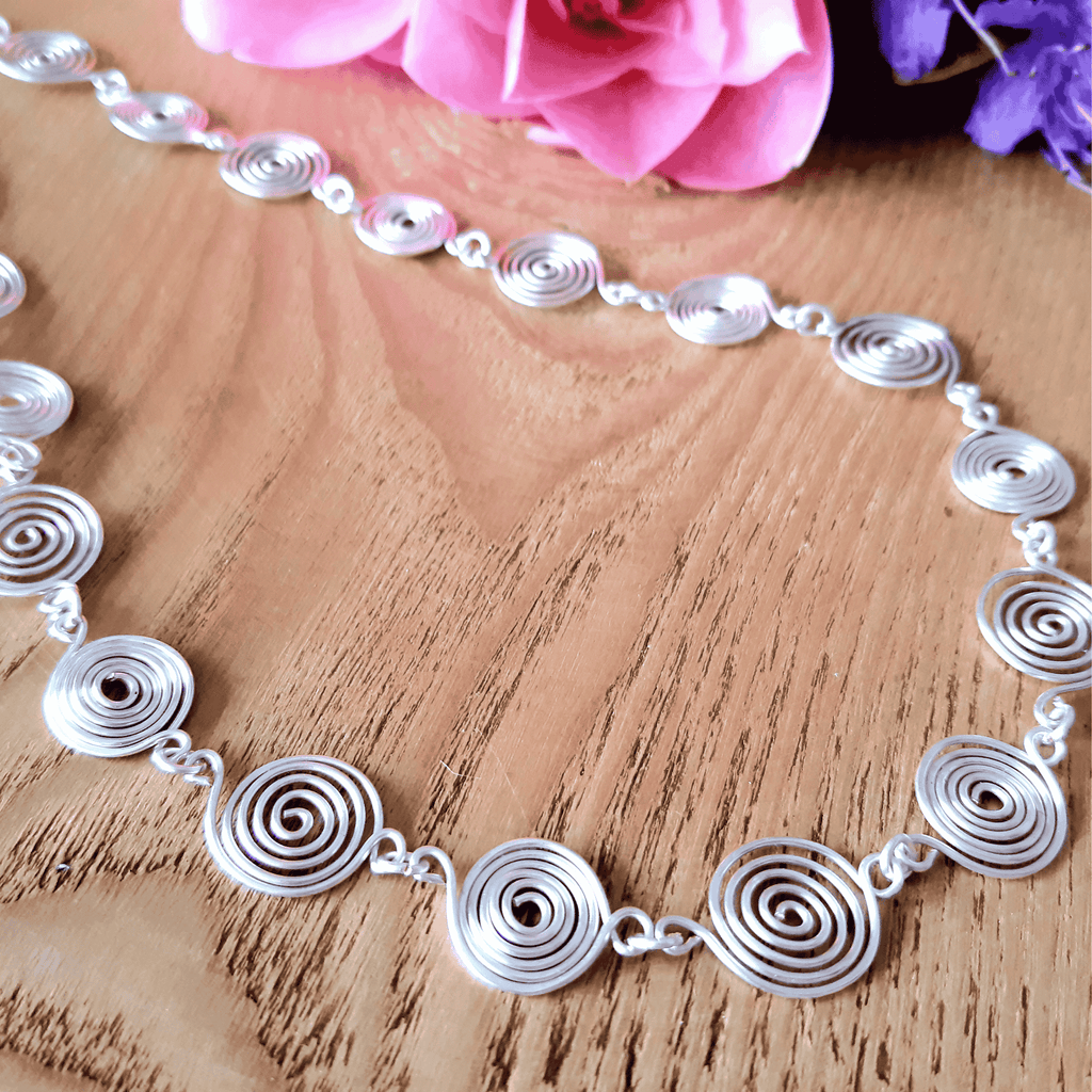Silver spiral necklace on a wooden surface with pink flowers in the background